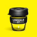 Glass keep cup with black lid and Lonsdale Roasters band, on brand yellow — Lonsdale Street Roasters